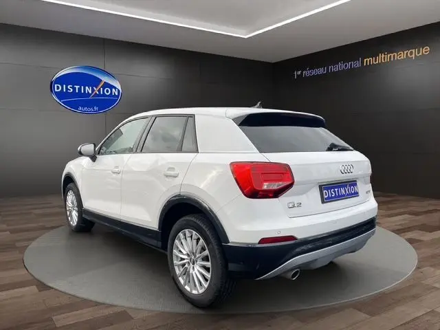 Audi Q2 blanc vue 3/4 arrière droit en intérieur, avec jantes alliage et feux arrière rouges distinctifs.