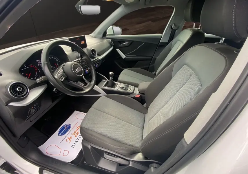 Vue intérieure côté conducteur de l'Audi Q2 30 TDI blanc, montrant les sièges tissu gris et le tableau de bord moderne.