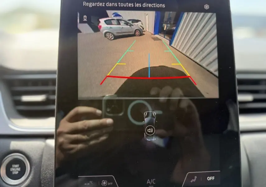 Vue écran caméra de recul montrant une Renault Captur gris Rafale avec toit noir, garée près d’un bâtiment industriel.