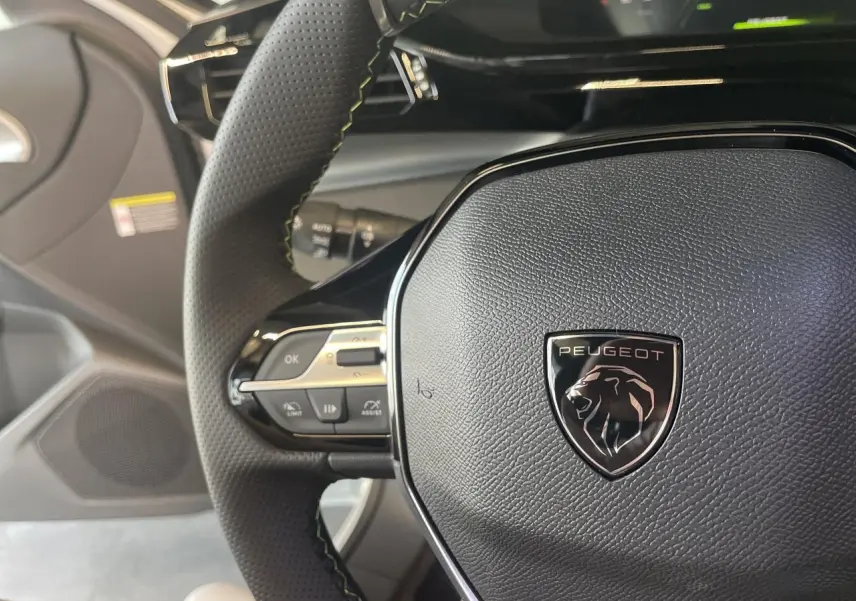 Gros plan sur le volant cuir noir perforé avec logo Peugeot et commandes intégrées dans une Peugeot 308 blanche 2025.
