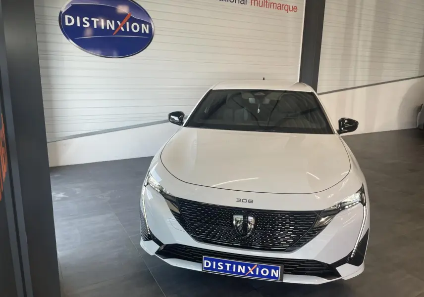 Vue avant d'une Peugeot 308 blanche 2025 avec calandre noire brillante et feux LED griffés, en showroom.