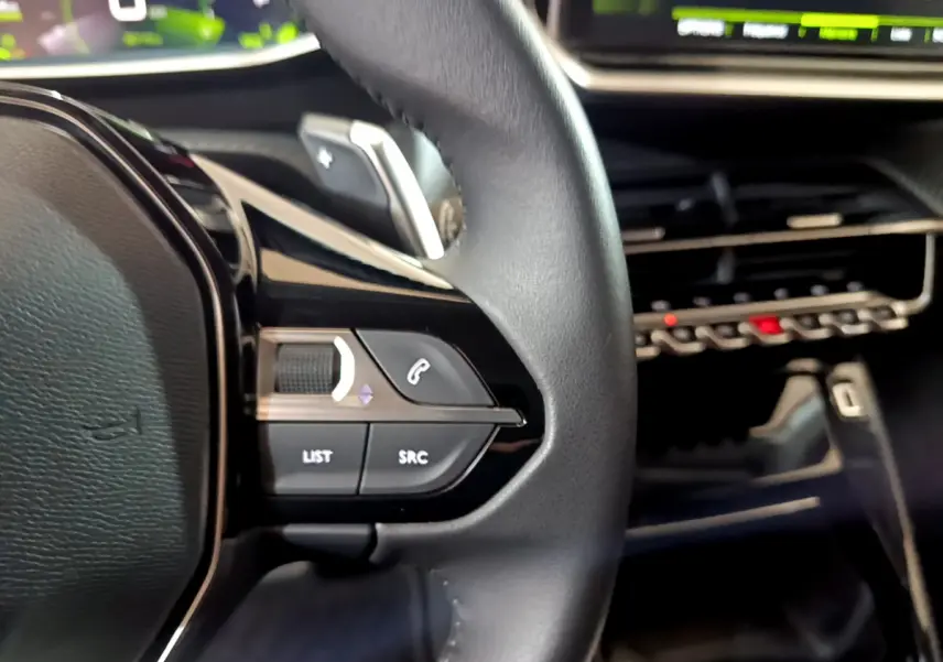 Gros plan sur les commandes gauche du volant cuir de la Peugeot 208 jaune 2023, avec tableau de bord numérique en arrière-plan.