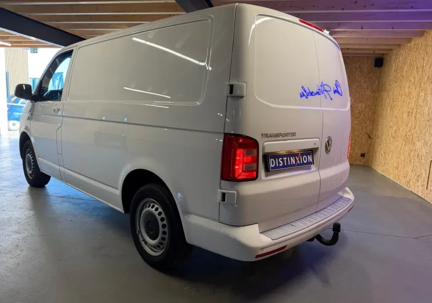 Vue 3/4 arrière droite d’un Volkswagen Transporter blanc 2019 en fourgon, avec attelage et feux arrière allumés.