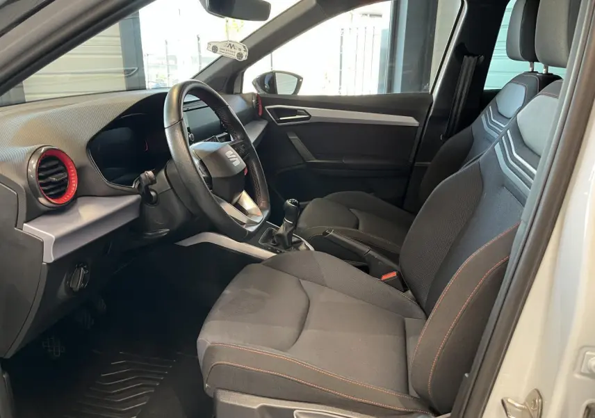 Intérieur côté conducteur du SEAT Arona blanc Nevada 2024, sièges textile noirs avec surpiqûres orange et volant sport cuir.