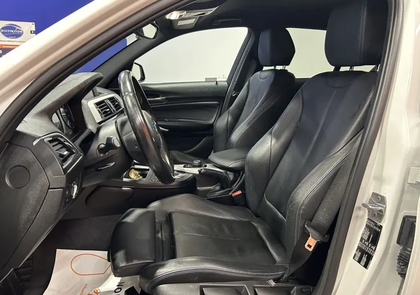 Vue intérieure côté conducteur d'une BMW Série 1 blanche de 2019, sièges en cuir noir avec coutures bleues et volant multifonction.