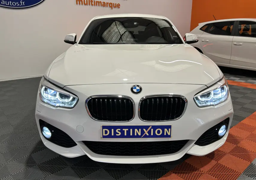Vue de face d'une BMW Série 1 blanche avec phares LED allumés et calandre noire brillante dans un showroom.
