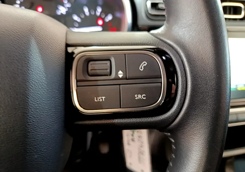 Gros plan sur les commandes du volant cuir côté gauche de la Citroën C3 blanche, avec écran tactile en arrière-plan.