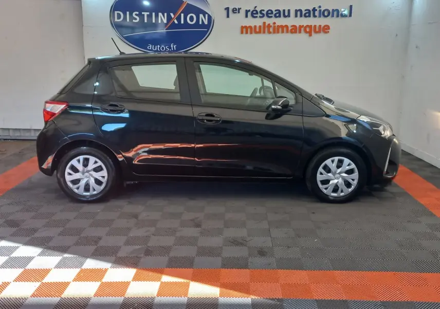 Profil côté gauche d'une Toyota Yaris noire 2018 hybride, stationnée en intérieur sur sol damier gris et orange.