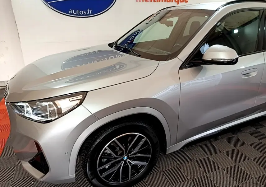 Vue 3/4 avant droit d’un BMW X1 xDrive23i M Sport gris clair avec jantes noires et phares LED allumés.