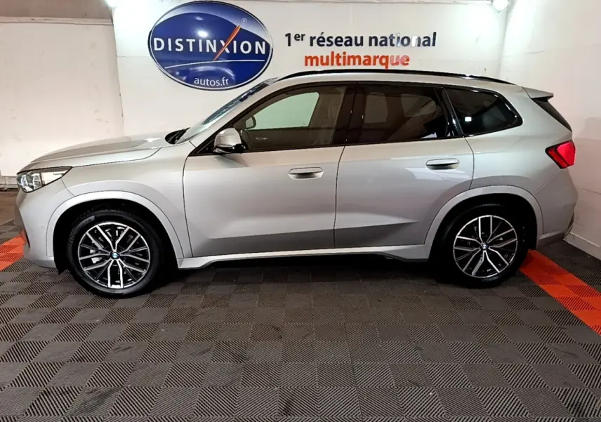 Vue de profil côté gauche d’un BMW X1 xDrive23i M Sport gris clair, roues alliage et vitres teintées, en intérieur showroom.
