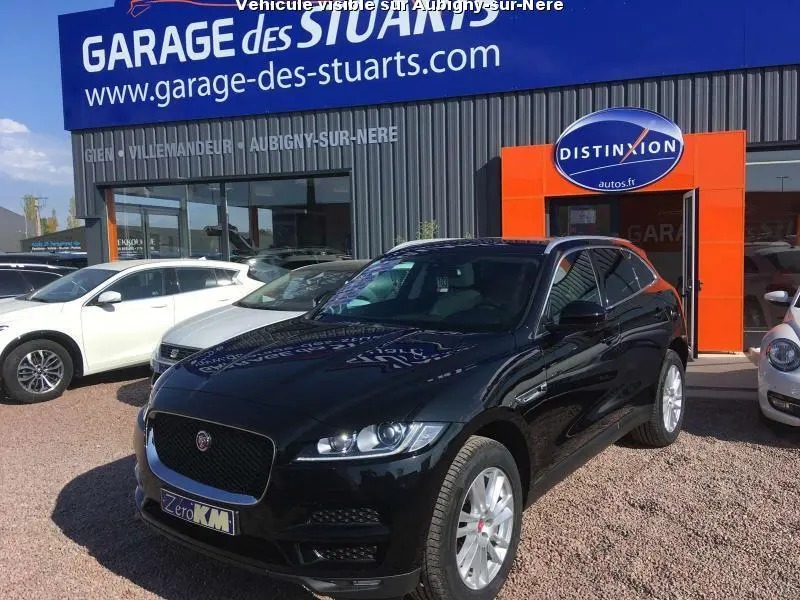 Jaguar F-PACE noir en 3/4 avant droit garé devant un garage avec jantes argentées et calandre noire.