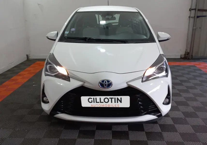 Vue avant d'une Toyota Yaris blanche 2019 avec phares allumés et calandre noire distinctive en intérieur showroom.