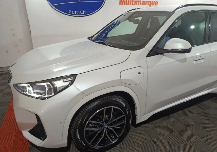 Vue 3/4 avant droit d’un BMW X1 blanc hybride rechargeable avec jantes noires et badge M Sport visible sur l’aile.