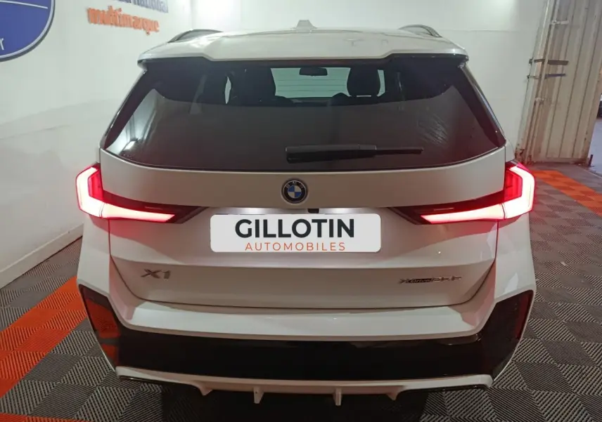 Vue arrière d’un BMW X1 blanc 2024 avec feux LED allumés et logo xDrive25e M Sport visible.