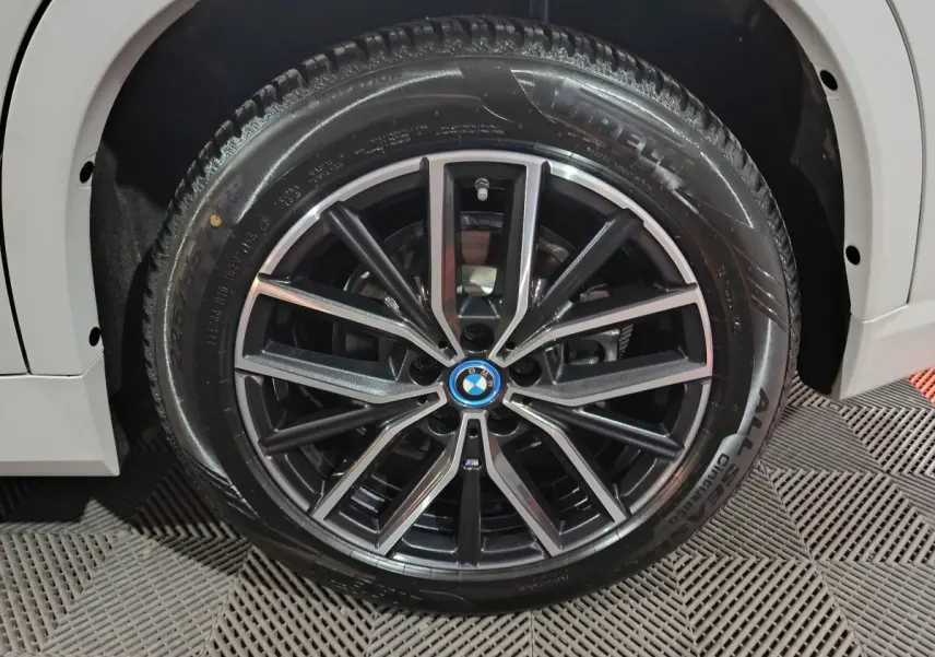 Gros plan sur la jante noire et argentée avec logo BMW d'un BMW X1 blanc, roue avant gauche visible.
