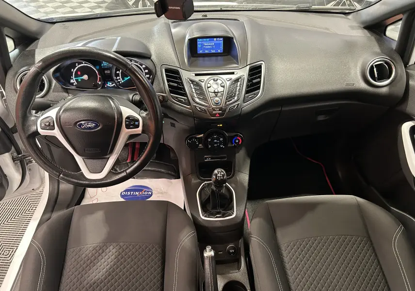 Vue intérieure avant de la Ford Fiesta blanche, montrant le tableau de bord, volant multifonction et levier de vitesse manuel.