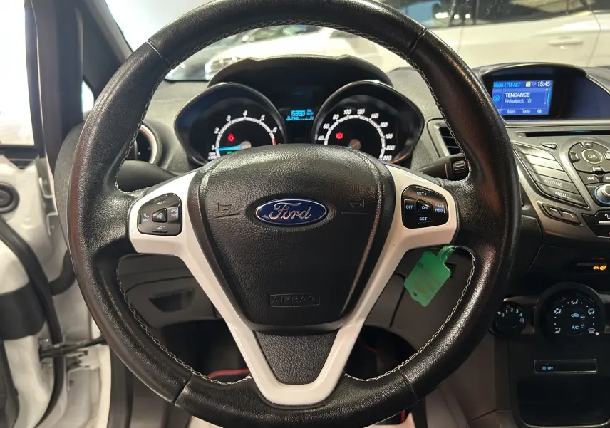 Vue rapprochée du volant en cuir noir avec commandes intégrées d’une Ford Fiesta blanche de 2015, tableau de bord visible.