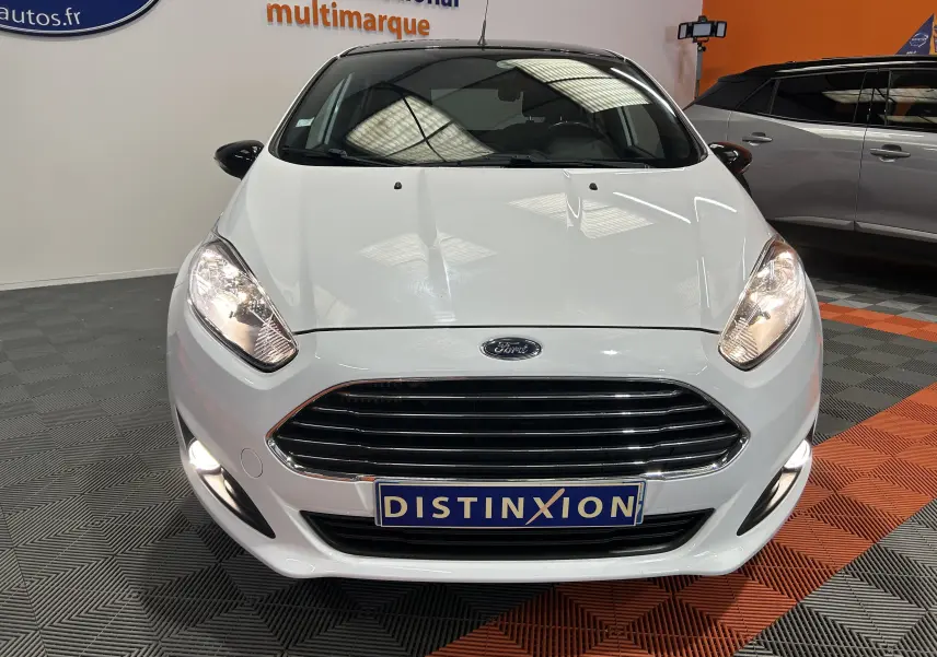 Vue avant d'une Ford Fiesta blanche EcoBoost 2015 avec phares allumés et calandre chromée dans un showroom.