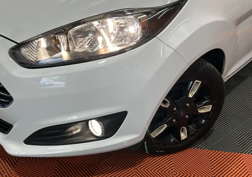 Gros plan sur l’avant droit blanc d’une Ford Fiesta 2015 avec phare et antibrouillard allumés, jante noire brillante.