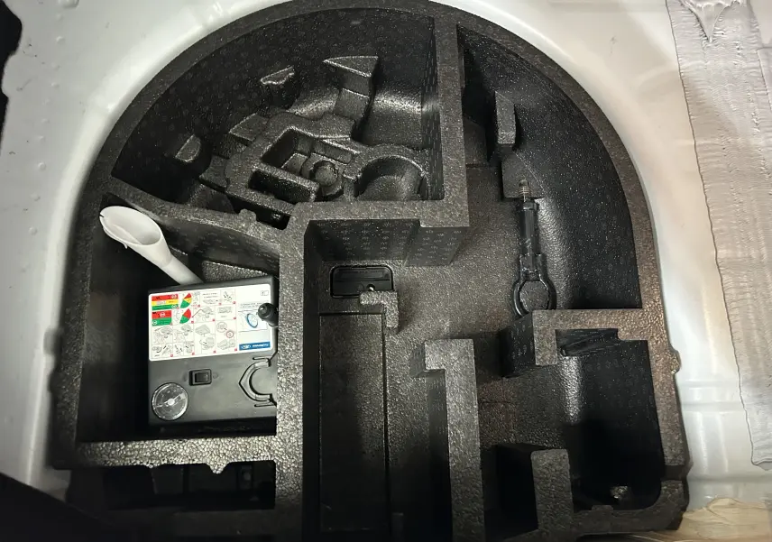 Vue en plongée sur le rangement sous coffre blanc d’une Ford Fiesta 2015, montrant le compresseur et un outil.