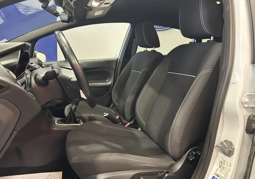 Vue intérieure côté gauche de la Ford Fiesta blanche 2015, montrant les sièges avant en tissu noir et le volant.
