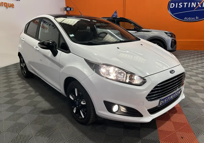 Vue 3/4 avant droite d'une Ford Fiesta blanche avec jantes noires et phares allumés dans un showroom.