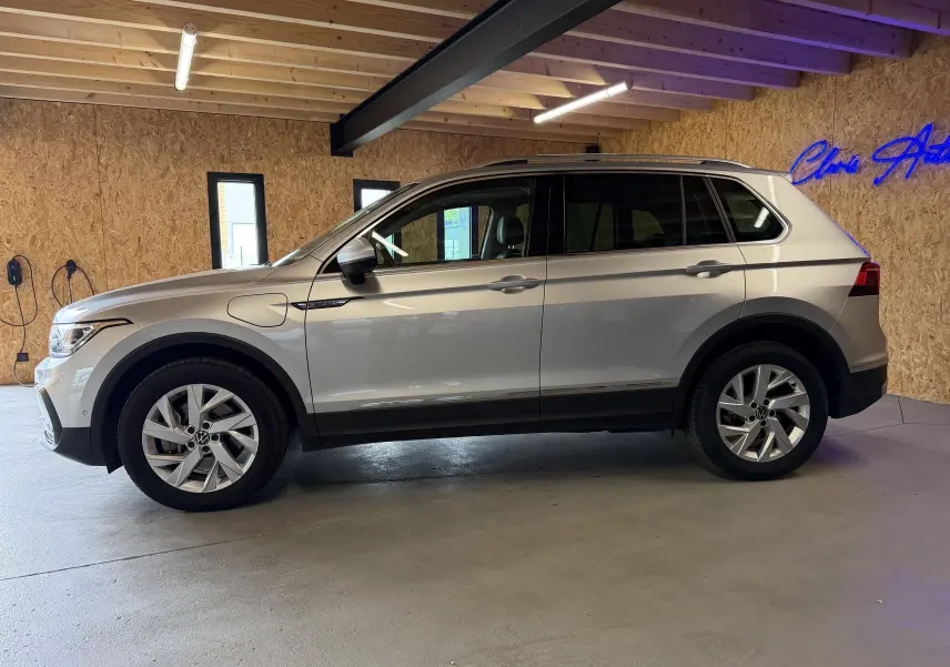 Vue de profil côté gauche d'un Volkswagen Tiguan 1.4 e-Hybrid gris clair, avec prises de recharge visibles sur l'aile avant.