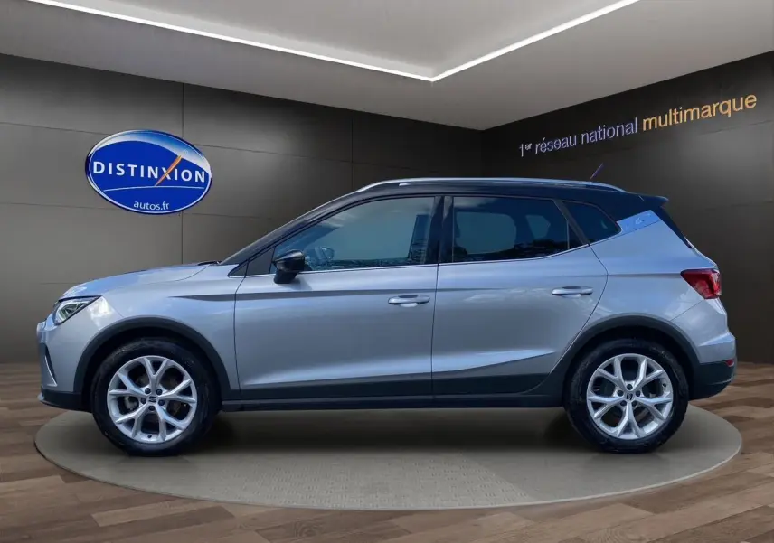 Vue de profil côté gauche d'un SEAT Arona gris silver avec toit noir et jantes alu 17 pouces dans un showroom.