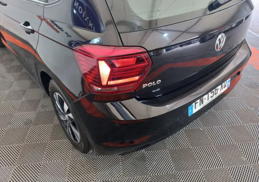 Vue 3/4 arrière gauche d'une Volkswagen Polo noire avec feux arrière allumés et logo visible sur hayon.