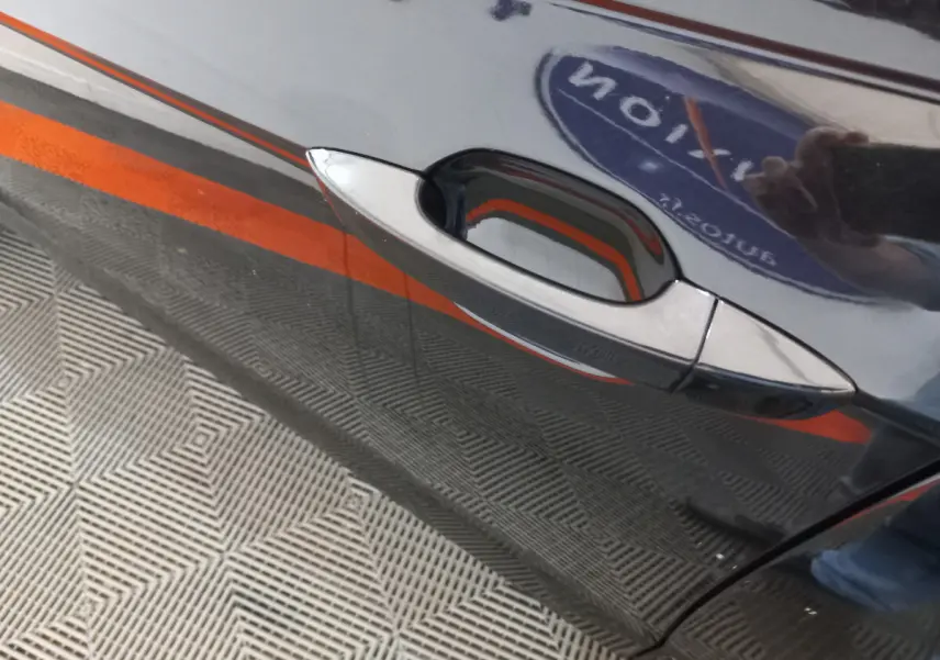 Poignée de porte côté gauche d'une Volkswagen Polo noire 2020 avec reflet d'un logo et sol à motifs géométriques.