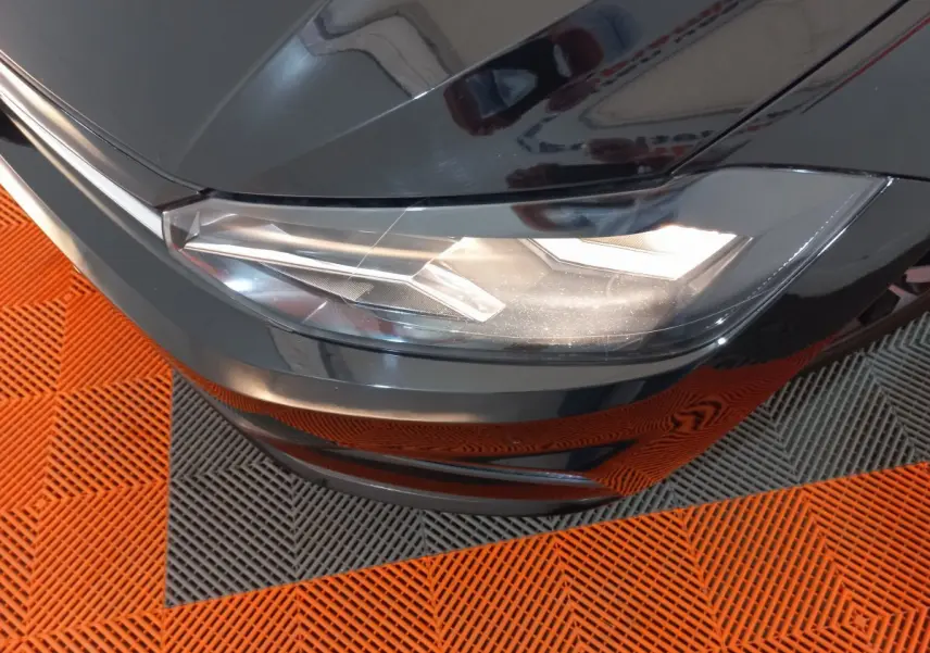 Gros plan sur le phare avant droit d'une Volkswagen Polo noire, avec reflets sur la carrosserie et sol orange et gris.