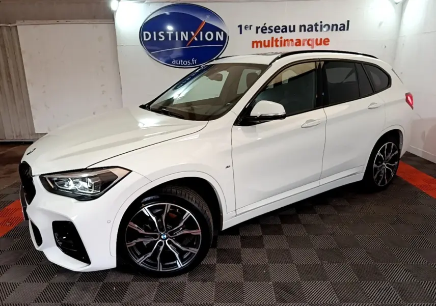 BMW X1 sDrive18i M Sport blanc vue 3/4 avant droit, jantes alliage noires et phares LED allumés, en intérieur showroom.