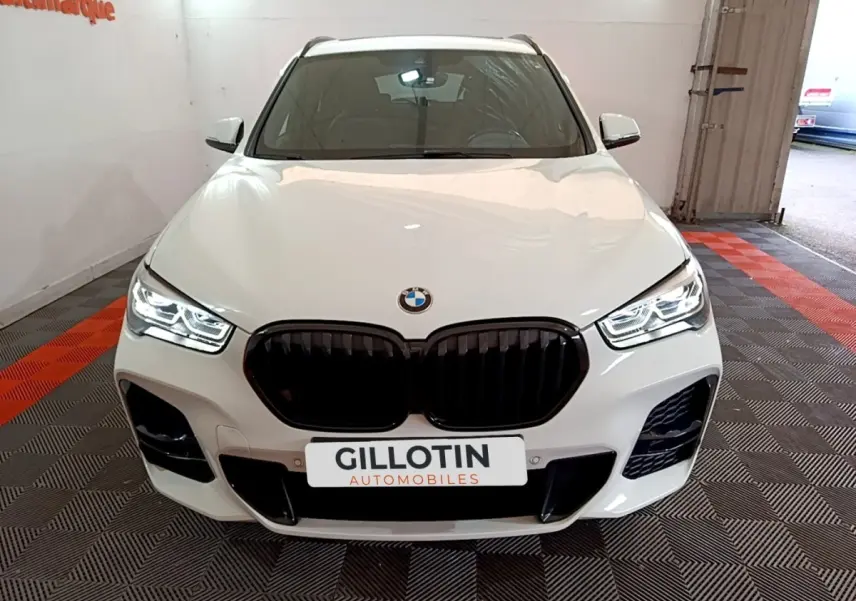 Vue avant d'une BMW X1 sDrive18i M Sport blanche avec calandre noire et phares LED allumés dans un showroom.