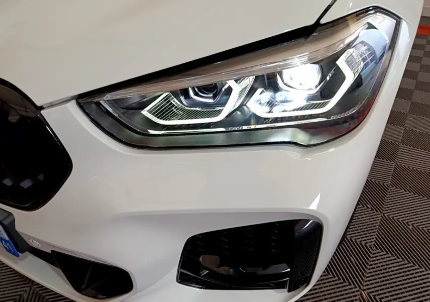 Gros plan sur le phare avant droit allumé d’un BMW X1 sDrive18i M Sport blanc, avec calandre noire partielle visible.