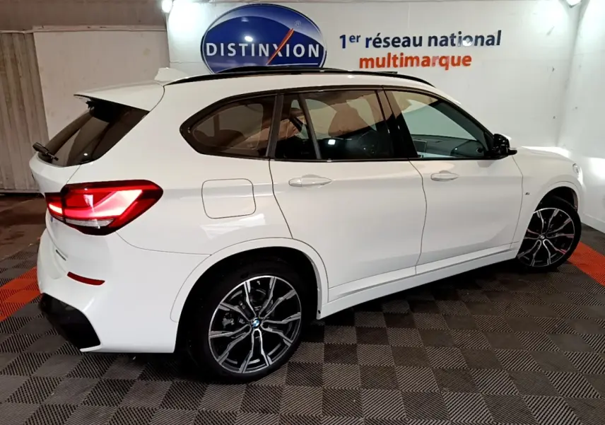 Vue 3/4 arrière droite d’un BMW X1 blanc sDrive18i M Sport avec jantes noires et feux arrière allumés.