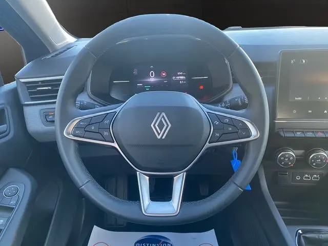 Vue rapprochée du volant textile noir avec logo Renault, tableau de bord numérique et commandes multifonctions du Renault Clio V gris schiste.