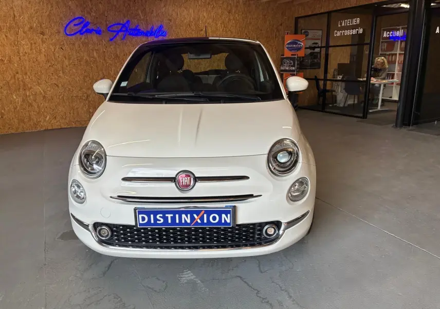 Vue de face d'une Fiat 500 blanc crème stationnée en intérieur, avec logo FIAT et plaque Distinxion visible.