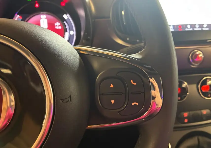 Gros plan sur le volant de la Fiat 500 blanc crème, avec commandes intégrées et tableau de bord lumineux en arrière-plan.