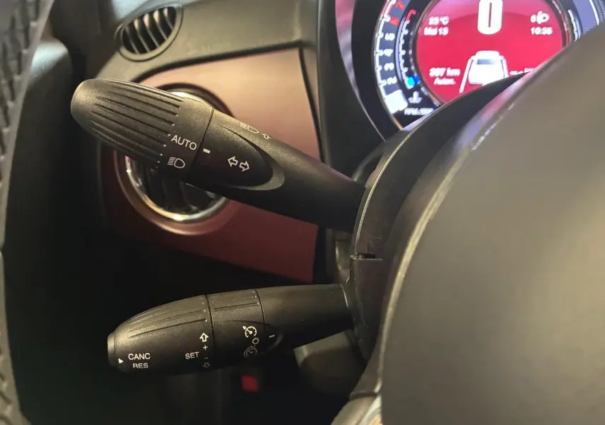 Gros plan sur les commandes de clignotants et régulateur de vitesse à gauche du volant d'une Fiat 500 blanc crème, tableau de bord numérique visible.