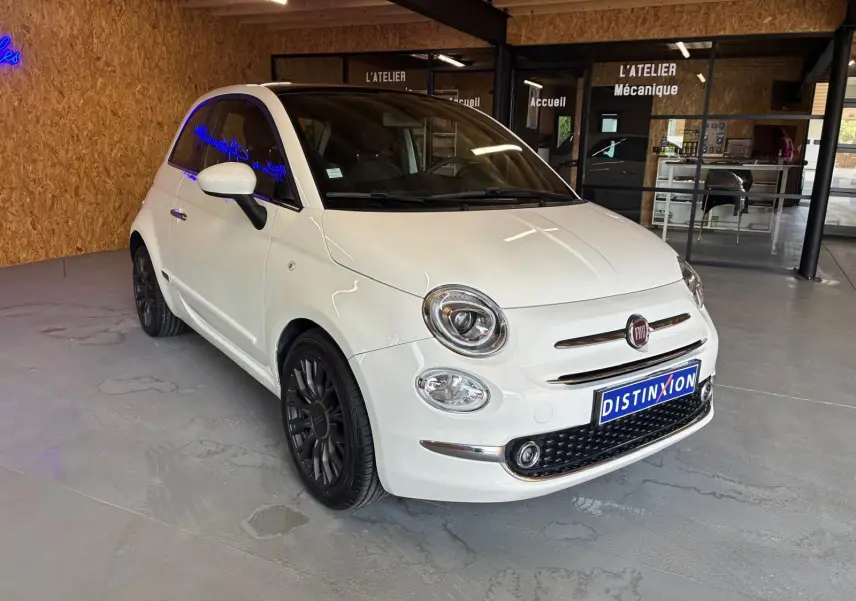 Vue 3/4 avant d'une FIAT 500 blanc crème, avec jantes noires et calandre chromée, stationnée en intérieur.