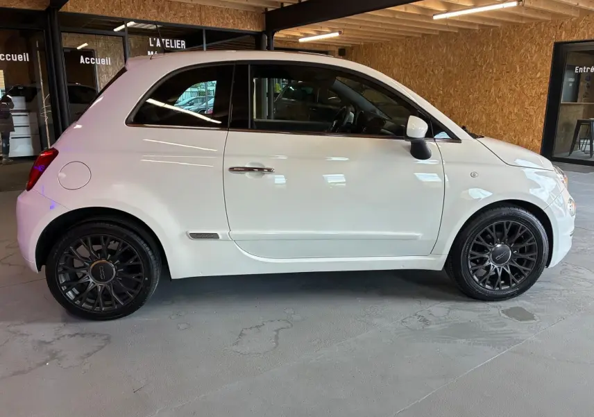 Vue de profil côté gauche d'une FIAT 500 blanc crème avec jantes noires en intérieur d'atelier.