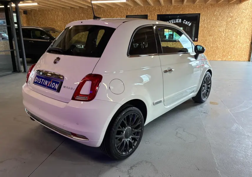 Vue 3/4 arrière droite d'une Fiat 500 blanc crème avec jantes noires et détails chromés dans un garage.