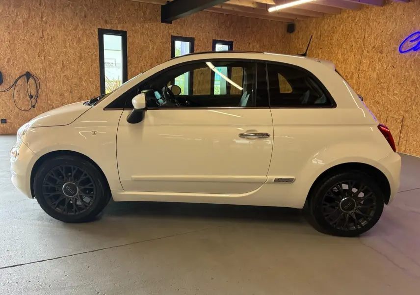 Vue de profil côté gauche d'une FIAT 500 blanc crème 2021 avec jantes noires et intérieur visible.
