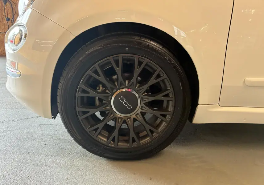 Gros plan sur la roue avant gauche noire à jante ajourée d'une FIAT 500 blanc crème vue de profil.