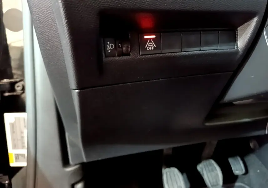 Gros plan sur les commandes sous le tableau de bord côté gauche du Peugeot 2008 noir, avec pédales et bouton d'aide à la conduite allumé.