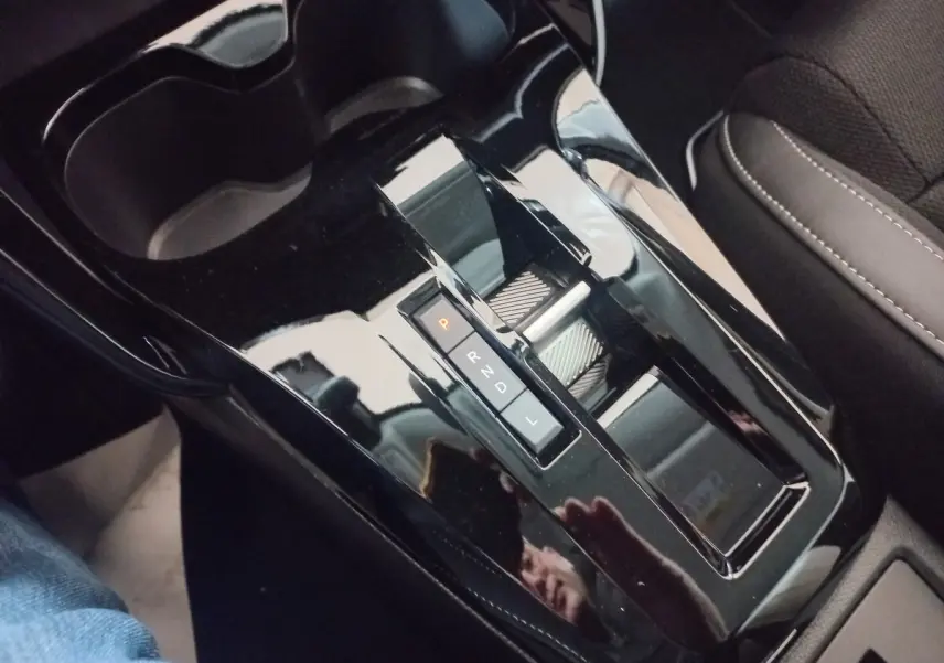 Gros plan sur la console centrale noire brillante avec levier de vitesses automatique et porte-gobelets, siège tissu gris visible.