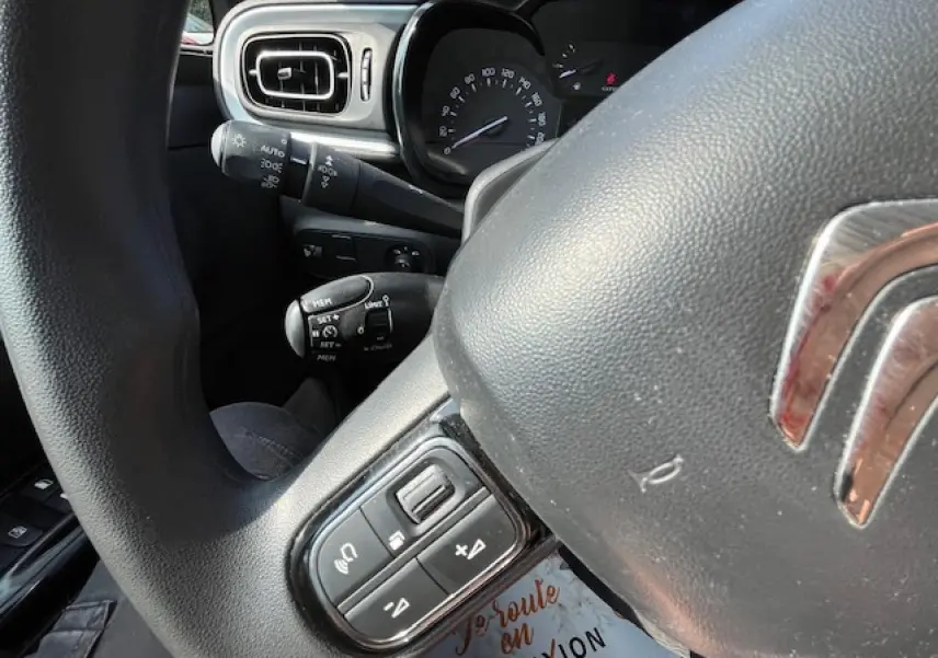 Gros plan sur le volant et le tableau de bord noir d'une Citroën C3 2023, avec commandes audio au volant visibles.