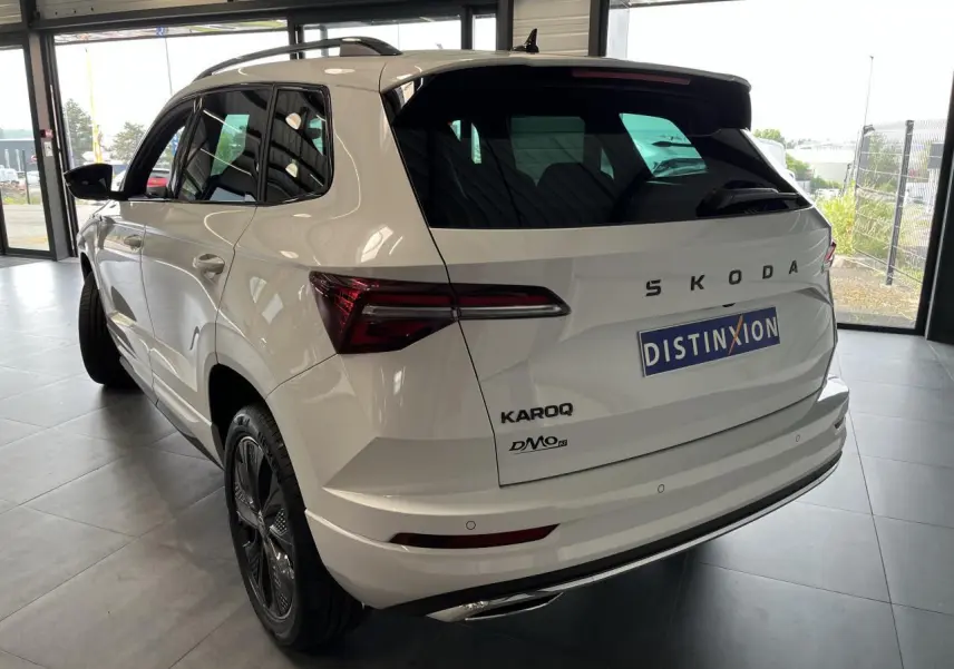 Vue 3/4 arrière droite du SUV blanc SKODA KAROQ 2025 avec jantes anthracite et hayon électrique en intérieur showroom.