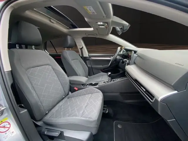 Intérieur avant de la Volkswagen Golf VIII gris argent, sièges tissu noir et toit panoramique ouvrant visible depuis la porte ouverte côté conducteur.