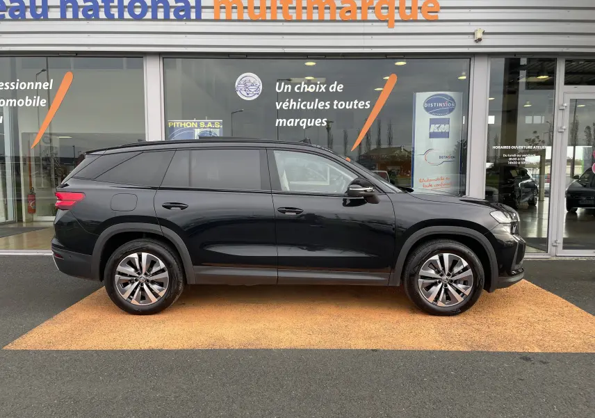 Profil latéral droit d'un SUV Skoda Kodiaq noir métal 7 places, garé devant une concession multimarque.